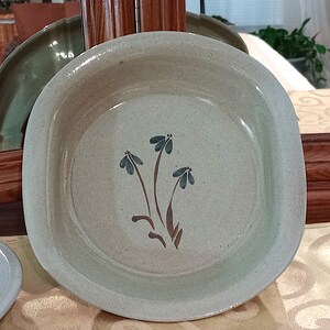 May include: A round, speckled, light beige stoneware bowl featuring a hand-painted design of three blue flowers with brown stems and leaves centered in the basin. The bowl has a smooth, glazed finish and a slightly raised rim, showcasing a simple, functional design.