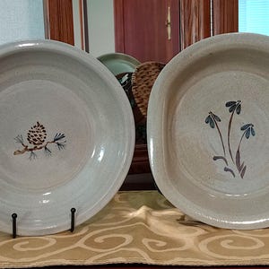 May include: Two ceramic bowls with different designs. One bowl has a pine cone and branch design, the other has a stylized flower design. Both bowls are light gray with a white glaze.