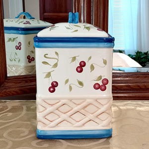 May include: White ceramic canister with a blue and white lid. The canister has a woven pattern on the bottom and a cherry design on the sides.