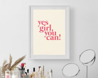 May include: A framed print with the text "yes girl, you can!" in pink lettering hangs above a white vanity. The vanity holds makeup, brushes, mirrors, and decorative items. A gold wire basket and dried plants add to the decor.
