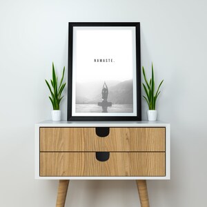 May include: A black framed print with the word "NAMASTE." in white text. The print features a black and white photo of a person doing yoga in front of a mountain range. The print is sitting on a wooden nightstand with two drawers and two legs. There are two green plants in white pots on either side of the print.