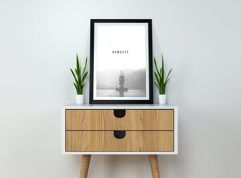 May include: A black framed print with the word "NAMASTE." in white text. The print features a black and white photo of a person doing yoga in front of a mountain range. The print is sitting on a wooden nightstand with two drawers and two legs. There are two green plants in white pots on either side of the print.