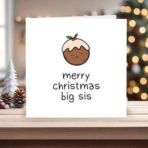May include: A white Christmas card featuring a cartoon Christmas pudding and the words "merry christmas big sis". The card is on a wooden surface, with a Christmas tree and festive decorations in the background.