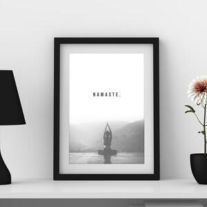 May include: A black and white framed print with the word "NAMASTE." printed in the center. The print features a silhouette of a person in a yoga pose in front of a mountain range. The print is on a white shelf with a black lamp and a potted white flower.