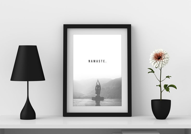 May include: A black and white framed print with the word "NAMASTE." printed in the center. The print features a silhouette of a person in a yoga pose in front of a mountain range. The print is on a white shelf with a black lamp and a potted white flower.