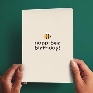 May include: A cream-colored greeting card featuring a small, illustrated yellow and black bee above the dark brown text that reads "happ-bee birthday!" with an exclamation point. The card is rectangular and oriented vertically.
