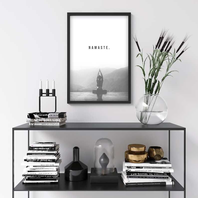 May include: A black and white framed print with the word "NAMASTE." printed in white on a white background. The print shows a person doing yoga in a lake. The print is on a black shelf with a candle holder, books, and decorative objects.