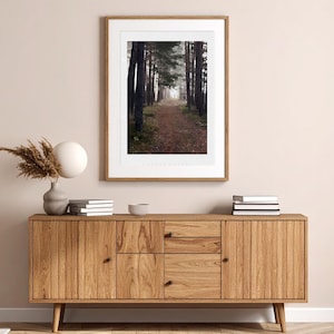 May include: A framed vertical wall art print featuring a forest path surrounded by tall, dark trees in a misty atmosphere. The print is displayed in a light wood frame hanging above a wooden sideboard with two cabinets and two drawers.