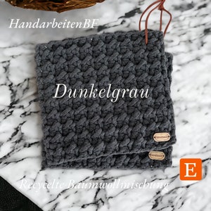 Könnte beinhalten: Zwei dunkelgraue gehäkelte Topflappen mit einem "Handmade"-Etikett auf jedem. Die Topflappen sind aus recycelter Baumwollmischung gefertigt und auf einer weißen Marmorfläche abgelegt.