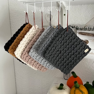 Könnte beinhalten: Ein Set aus sechs gehäkelten Topflappen in verschiedenen Farben, darunter Schwarz, Braun, Weiß, Beige, Grau und Dunkelgrau. Die Topflappen hängen an Haken.