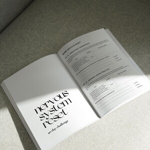 May include: An open book with the title "nervous system reset" and the subtitle "40-day challenge" in a modern font. The pages are white with black text and a section titled "Day 15 - Predictable Soundtrack". The book is on a light gray surface.