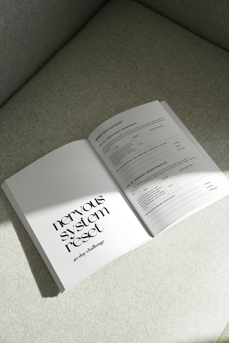 May include: An open book with the title "nervous system reset" and the subtitle "40-day challenge" in a modern font. The pages are white with black text and a section titled "Day 15 - Predictable Soundtrack". The book is on a light gray surface.