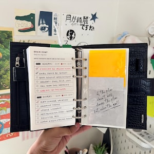 May include: A black crocodile-textured leather ring binder planner featuring a six-ring metal mechanism. The open planner displays a white paper insert titled Brain Dump with handwritten task lists and a translucent page with the text If the plan.