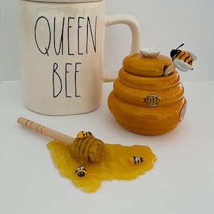 May include: White ceramic mug with a bee lid and the text "Queen Bee". A yellow ceramic honey pot shaped like a beehive with a bee on the lid and a wooden honey dipper. The dipper is in a puddle of honey with several small bees.