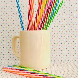 May include: A white ceramic mug filled with colorful striped drinking straws. The mug has a handle and is sitting on a white surface with a colorful polka dot pattern.