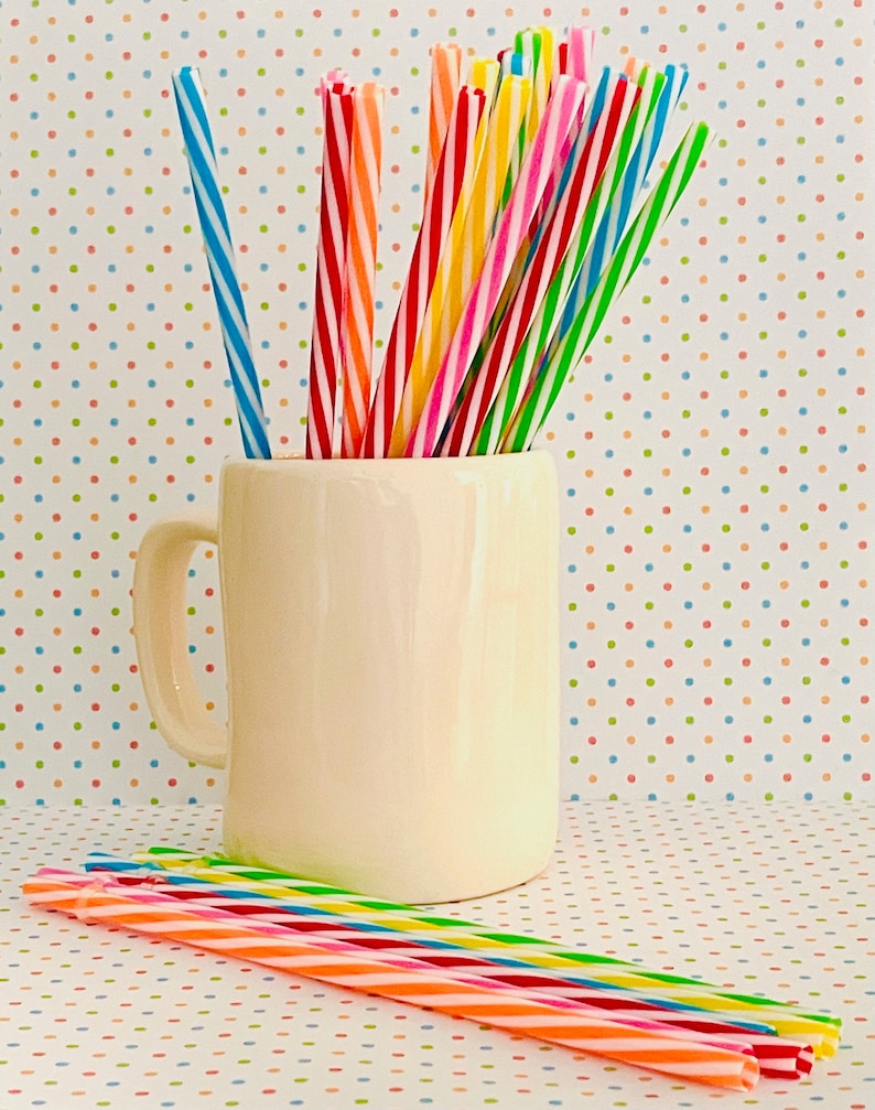 May include: A white ceramic mug filled with colorful striped drinking straws. The mug has a handle and is sitting on a white surface with a colorful polka dot pattern.