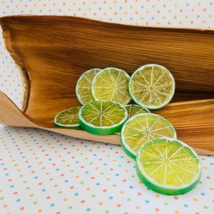 May include: A group of lime slices arranged on a brown, woven surface. The lime slices are green and yellow and have a slightly textured surface.