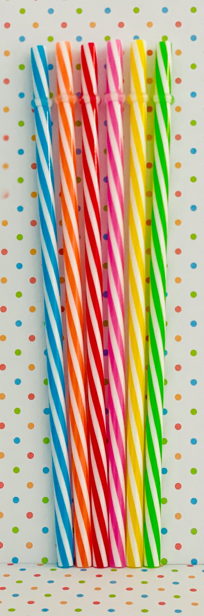May include: A set of seven striped drinking straws in various colors, including blue, orange, red, pink, yellow, and green. The straws are arranged vertically against a white background with colorful polka dots.