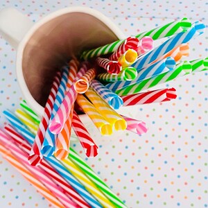 May include: A white ceramic mug filled with colorful striped drinking straws. The straws are arranged in a fan shape, with the ends of the straws sticking out of the mug. The mug is sitting on a white surface with a polka dot pattern.