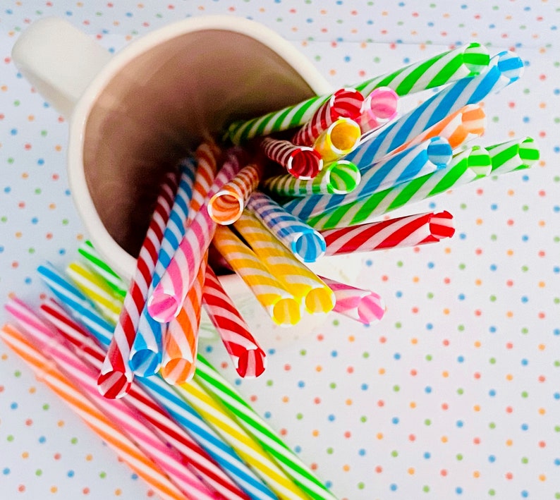 May include: A white ceramic mug filled with colorful striped drinking straws. The straws are arranged in a fan shape, with the ends of the straws sticking out of the mug. The mug is sitting on a white surface with a polka dot pattern.