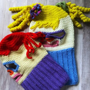 May include: Two crocheted hats with colorful stripes and a face design. One hat is yellow, blue, and pink with a long yellow yarn tail. The other hat is red, white, purple, and blue.