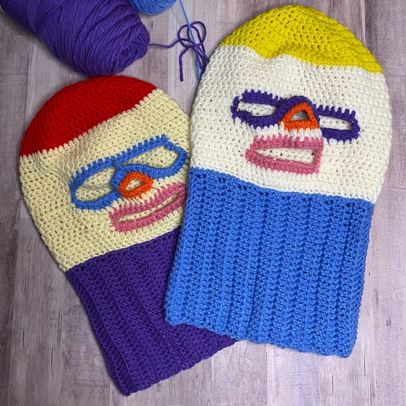 May include: Two crocheted balaclavas, one with a yellow, white, and blue color scheme and the other with a red, white, and purple color scheme. Both balaclavas have a crocheted face design with a mouth, eyes, and nose.