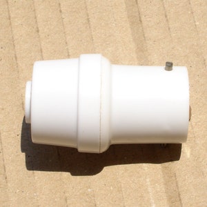 May include: A white plastic cylindrical object with a slightly textured surface. The object has a wider central section and narrower ends, with a small metal pin on one end. Likely a plumbing fitting or connector.