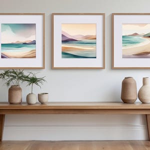 Könnte beinhalten: Drei gerahmte Aquarellbilder von Meereslandschaften mit Sandstränden, türkisfarbenem Wasser und fernen Hügeln. Die Bilder werden auf einem Holzregal mit zwei Vasen und einer Topfpflanze ausgestellt.