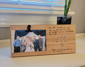 Puede incluir: Un marco de fotos de madera clara con un clip negro que sostiene una foto de boda. El marco tiene texto escrito a mano. Una pequeña jarra de cristal con tallos de bambú encima.