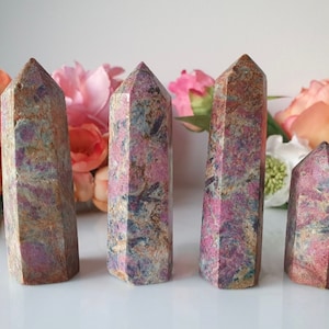 May include: Four pink, brown, and grey gemstone points, also known as ruby in fuchsite, are arranged in a row on a white surface. The points are polished and have a smooth, shiny finish.