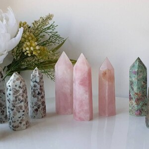 May include: A collection of polished crystal points in various colors, including green, pink, and grey. The points are arranged in a row on a white surface.