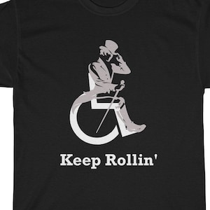 Puede incluir: Camiseta negra con una silueta blanca de una persona en silla de ruedas con un sombrero de copa y un bast&oacute;n. El texto "Keep Rollin'" est&aacute; impreso en blanco debajo de la imagen.