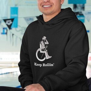 Puede incluir: Una persona con una sudadera con capucha negra con el texto "Keep Rollin'" y un gr&aacute;fico de una persona en silla de ruedas est&aacute; sentada en una silla de ruedas verde. La silla de ruedas est&aacute; frente a una piscina.