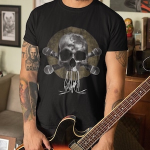 Può includere: T-shirt nero con un design a teschio e ossa incrociate e una chitarra. Il teschio è centrato su un cerchio giallo sbiadito. La chitarra ha una finitura marrone e arancione. La persona indossa un berretto nero.