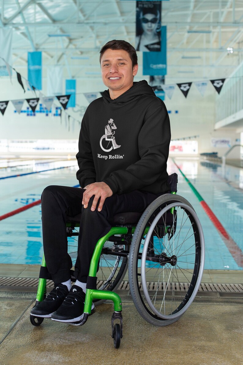 Puede incluir: Una persona con una sudadera con capucha negra con el texto "Keep Rollin'" y un gr&aacute;fico de una persona en silla de ruedas est&aacute; sentada en una silla de ruedas verde. La silla de ruedas est&aacute; frente a una piscina.