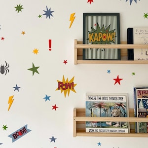 Puede incluir: Una pared blanca decorada con pegatinas de pared coloridas estilo cómic que incluyen estrellas, arañas, rayos y signos de exclamación, junto a burbujas de texto que dicen WHAM, KAPOW, POW y BOOM, con dos estantes de madera que sostienen libros.