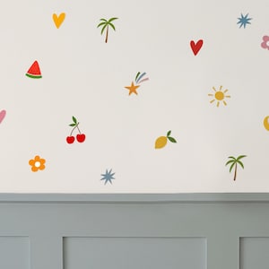 Op de afbeelding: Een witte muur versierd met kleurrijke, grillige stickers. De stickers bevatten harten, kersen, bloemen, sterren, palmbomen, een watermeloen, een citroen en een zon. De muur bevindt zich boven een grijs paneel.