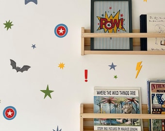 May include: A white wall decorated with colorful superhero-themed decals, including stars, lightning bolts, bats, and masks. Two wooden shelves hold framed art and children's books, creating a playful and imaginative space.