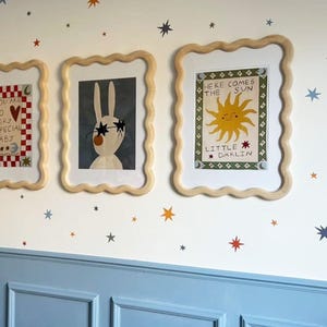 Puede incluir: Tres impresiones enmarcadas con ilustraciones caprichosas en una pared blanca con zócalo azul. Las impresiones muestran un conejo con gafas de sol, un sol con una cara sonriente y un patrón a cuadros con el texto "You are so very special baby". La pared está decorada con pequeñas pegatinas de estrellas en varios colores.