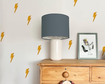 May include: A white wall adorned with yellow lightning bolt decals. A wooden dresser holds a white lamp with a gray shade, and a framed illustration. A plaid shirt hangs below. Home decor.