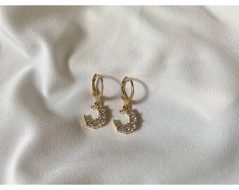 May include: A pair of gold hoop earrings with a crescent moon charm adorned with clear cubic zirconia stones.