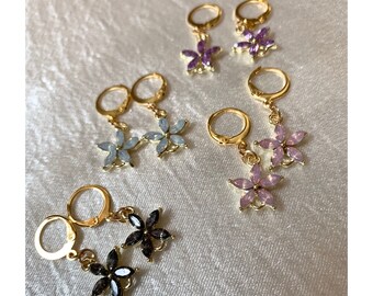 May include: A set of six pairs of gold-tone dangle earrings with flower-shaped charms. The charms are adorned with rhinestones in various colors, including purple, light blue, and black.