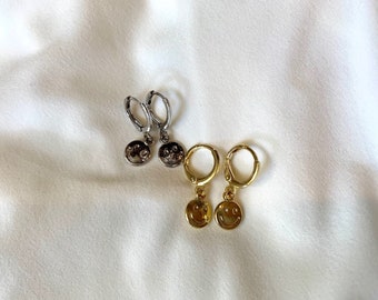 May include: Two pairs of hoop earrings. One pair is silver-toned, and the other is gold-toned. Each earring features a small, round charm with a smiling face design. The earrings are displayed on a white, textured surface.