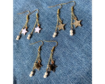May include: Three pairs of dangle earrings with star charms. The first pair has white enamel stars, the second pair has sparkly gold stars, and the third pair has black enamel stars. Each pair has a small pearl bead on the chain.
