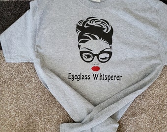 May include: Gray t-shirt with a black and white illustration of a woman wearing glasses and a red lipstick. The text "Eyeglass Whisperer" is printed below the illustration.