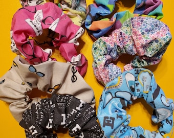 May include: Six colorful scrunchies made of fabric with different patterns. The scrunchies are in a variety of colors, including pink, blue, green, yellow, and black. The patterns include cats, eyeglasses, and abstract designs.