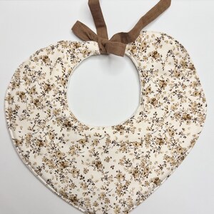 May include: Heart-shaped baby bib with a cream background and a pattern of small brown flowers. The bib has a brown fabric tie at the top for securing around the neck. The bib is made of soft fabric.