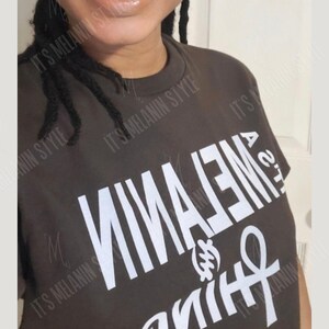 May include: A dark brown t-shirt with the words "It's Melanin" printed in white, with the word "Melanin" upside down. The shirt also features a white Ankh symbol.