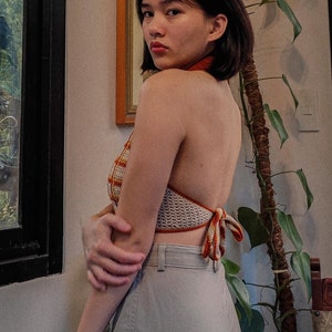 May include: A woman wearing a crocheted halter top with orange and white stripes. The top is tied at the back and has a loose fit. The woman is wearing light gray pants.