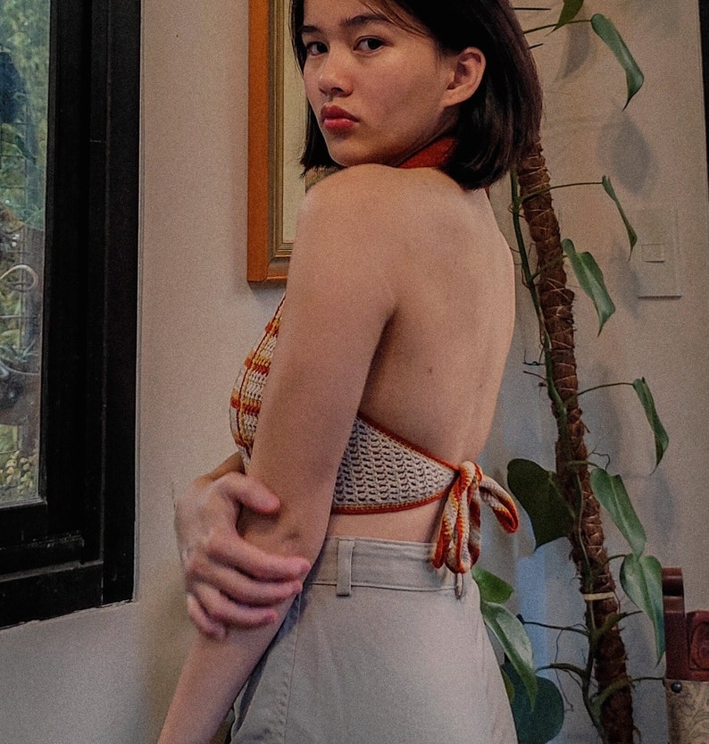 May include: A woman wearing a crocheted halter top with orange and white stripes. The top is tied at the back and has a loose fit. The woman is wearing light gray pants.